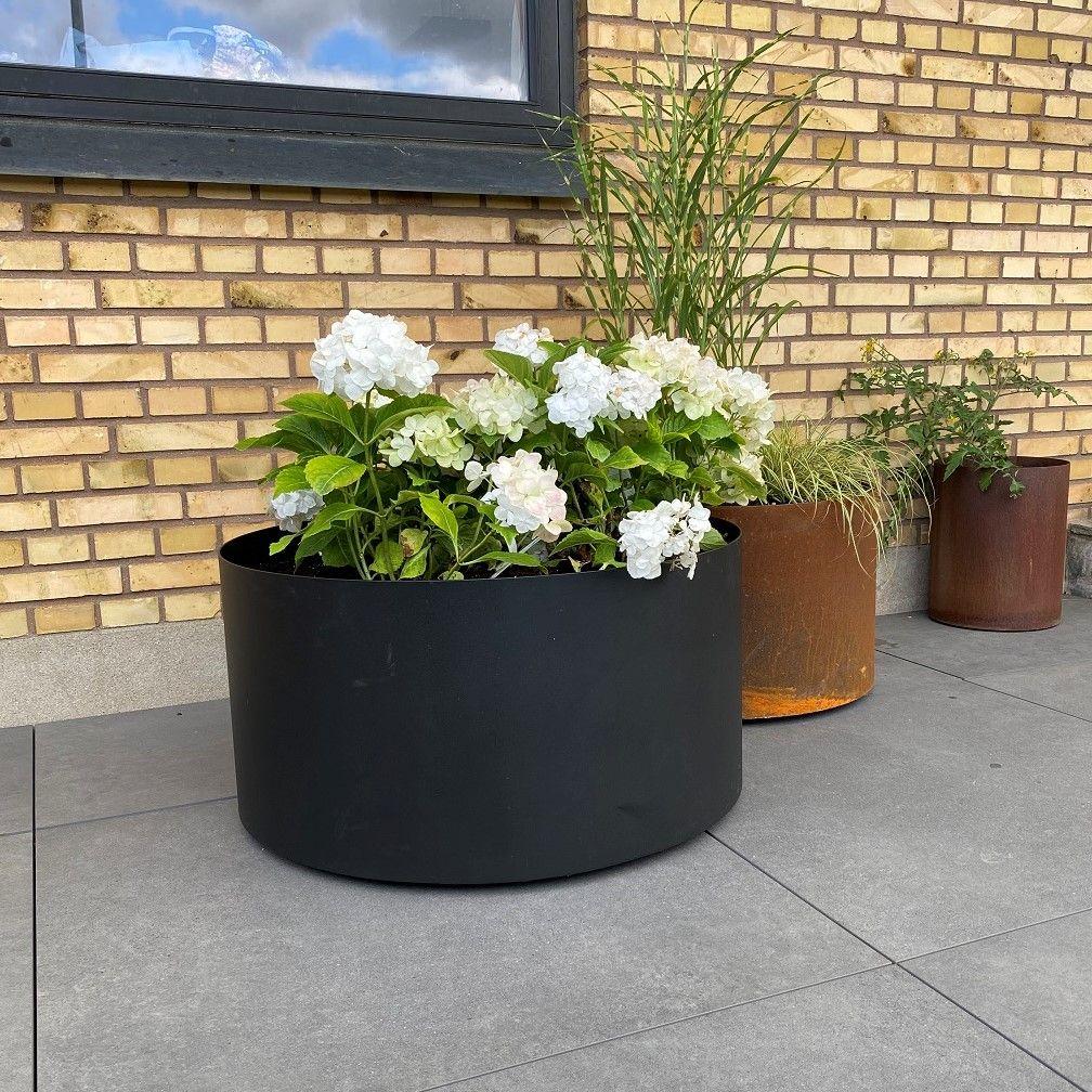 Stort rundt højbed i sort stål med hvid hortensia i forgrunden og to mindre runde højbede i cortenstål bagved op ad en murstensvæg