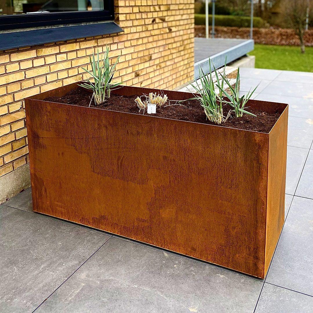 Højt, rektangulært højbed i cortenstål med grønne planter, placeret op ad en murstensfacade på en flisebelagt terrasse